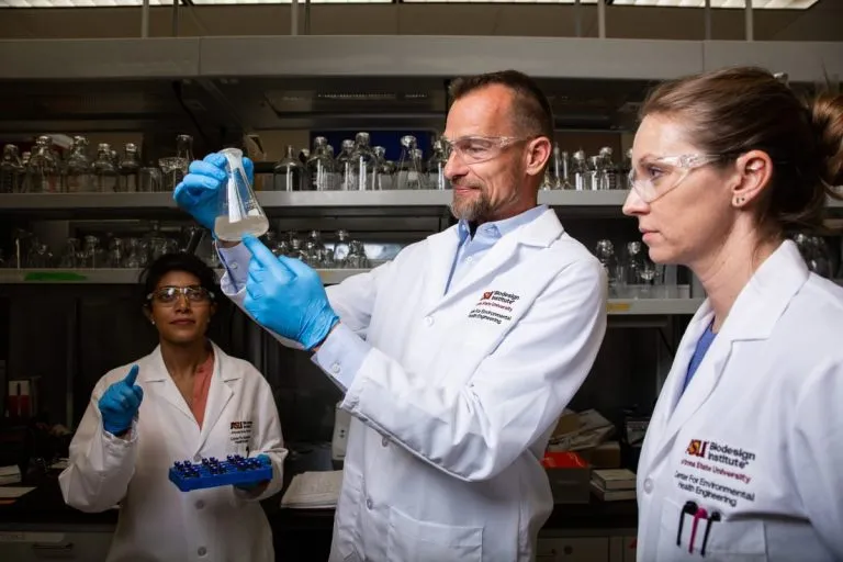 three people dressed in white coats, with blue gloves and safety glasses, one of them checks a container of cloudy water