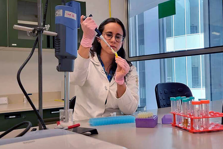 Middle aged woman, long hair, with glasses and wearing a white coat and latex gloves, is handling scientific instruments inside a laboratory