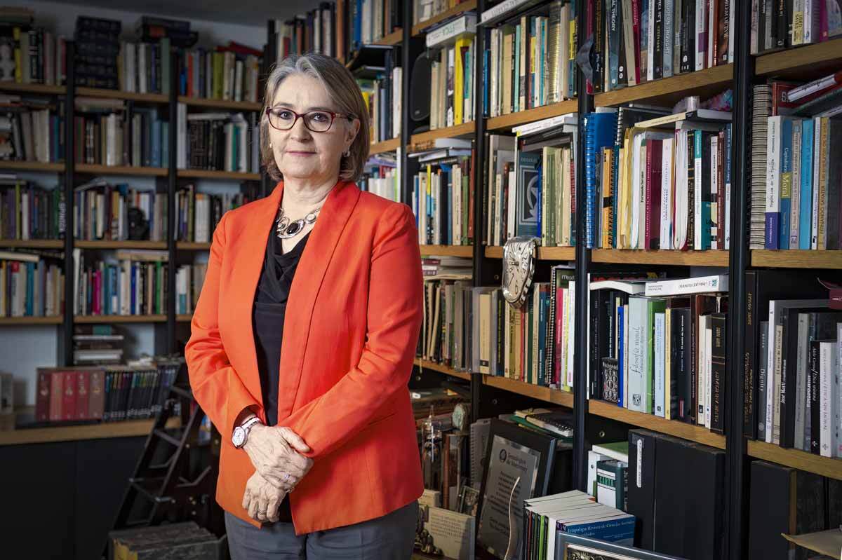 Middle-aged woman with an orange jacket and glasses in front of a bookshelf.