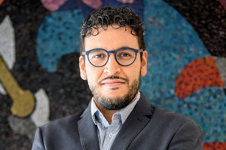 Man with glasses, a beard and dressed in a suit posses in front of a colorful mural