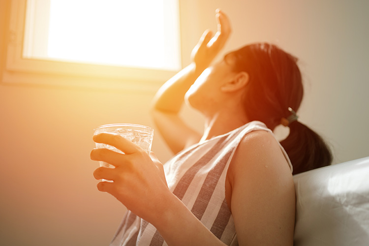 The photograph shows a person sitting and leaning against a pillow with sunlight streaming through a window. They are holding a transparent glass with a beverage, possibly water, and seem to be in the process of drinking or about to drink.