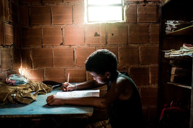 Image of a dimly lit room made of bricks, where a girl is seen writing in a notebook.