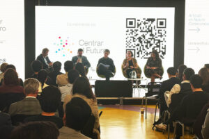 A panel of five people seated on a stage facing an audience, with a screen in the background displaying the "Centrar el Futuro" logo and a QR code.