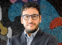 Man with glasses, a beard and dressed in a suit posses in front of a colorful mural