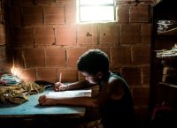 Image of a dimly lit room made of bricks, where a girl is seen writing in a notebook.