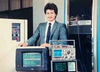 man in gray suit and blue tie surrounded by 80s computer equipment