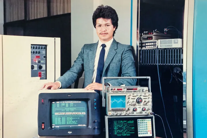 hombre con traje gris y corbata azul rodeado de equipos de cómputo de los años 80