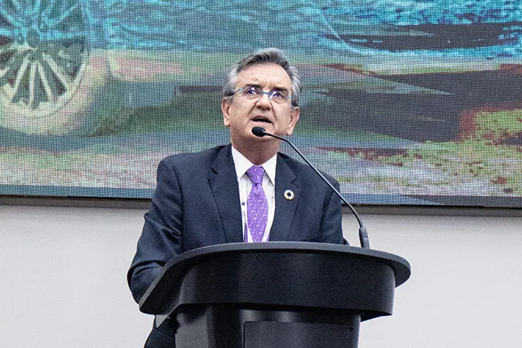 Hombre canoso, vistiendo un traje oscuro y corbata roja, haciendo una presentación