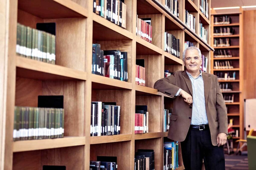 Hombre vestido con saco recargado en un librero de doble altura en una biblioteca