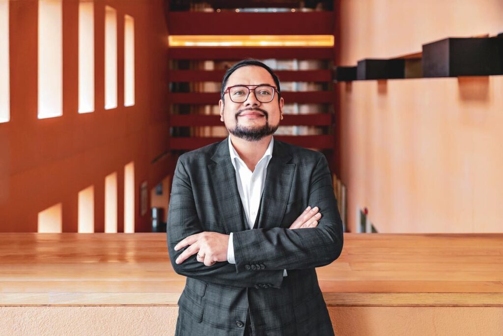 Hombre de mediana edad, con bigote y lentes, con traje azul cruzado de brazos.