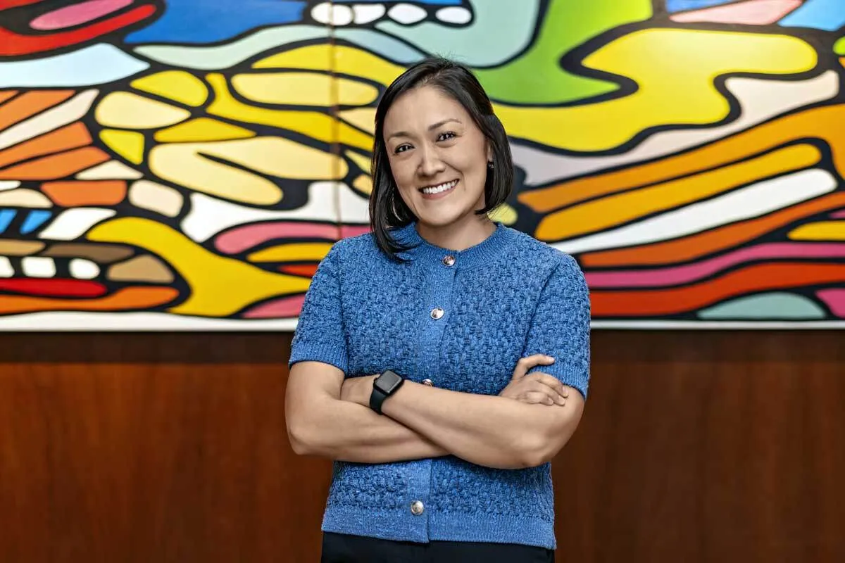 Mujer sonriendo con los brazos cruzados. Trae una blusa azul y está frente a un cuadro muy colorido