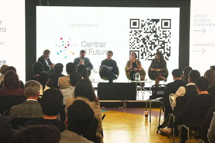 Panel de cinco personas sentadas en un escenario frente a una audiencia, con una pantalla de fondo que muestra el logo "Centrar el Futuro" y un código QR.
