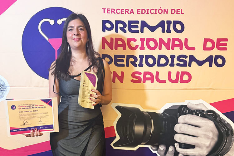 Inés Gutiérrez Jaber sonríe mientras sostiene el trofeo y el diploma del Tercer Premio Nacional de Periodismo en Salud 2026, con una vista nocturna de la ciudad de fondo.