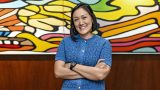 Mujer sonriendo con los brazos cruzados. Trae una blusa azul y está frente a un cuadro muy colorido