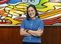 Mujer sonriendo con los brazos cruzados. Trae una blusa azul y está frente a un cuadro muy colorido