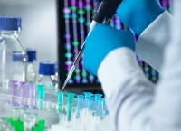 La imagen muestra un entorno de laboratorio en primer plano, donde una persona con bata blanca y guantes azules utiliza una pipeta para transferir un líquido a uno de varios tubos de ensayo pequeños. Los tubos de ensayo están dispuestos en un soporte y contienen líquidos de varios colores, incluyendo azul, verde y transparente. En el fondo, se ven imágenes borrosas de otros equipos de laboratorio y gráficos de barras verticales en una pantalla de computadora o monitor. Esta imagen representa investigaciones científicas o experimentos realizados en un entorno de laboratorio, relacionados con campos como la biología, la química o la medicina.