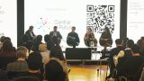 Panel de cinco personas sentadas en un escenario frente a una audiencia, con una pantalla de fondo que muestra el logo "Centrar el Futuro" y un código QR.