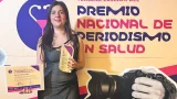 Inés Gutiérrez Jaber sonríe mientras sostiene el trofeo y el diploma del Tercer Premio Nacional de Periodismo en Salud 2026, con una vista nocturna de la ciudad de fondo.