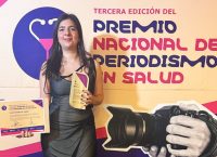 Inés Gutiérrez Jaber sonríe mientras sostiene el trofeo y el diploma del Tercer Premio Nacional de Periodismo en Salud 2026, con una vista nocturna de la ciudad de fondo.