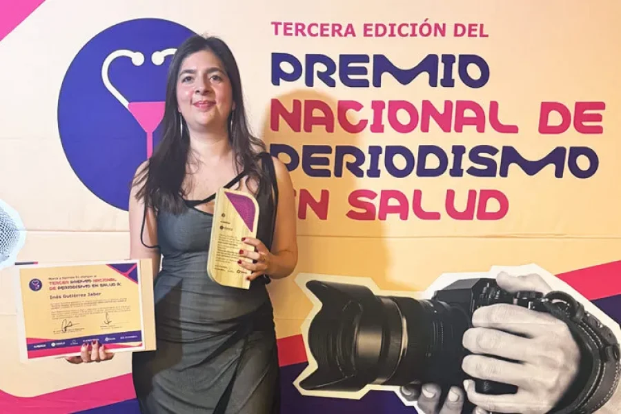 Inés Gutiérrez Jaber sonríe mientras sostiene el trofeo y el diploma del Tercer Premio Nacional de Periodismo en Salud 2026, con una vista nocturna de la ciudad de fondo.