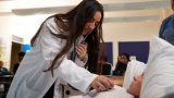Mujer con cabello largo y un estestoscopio, vistiendo bata blanca, colocando el estetoscopio sobre un paciente