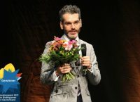 Hombre joven con lentes, traje gris y sosteniendo con una mano una flores y con la otra mano sosteniendo un microfono